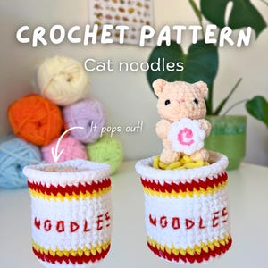 Puede incluir: Patrón de crochet "Cat Noodles" con dos tazas de fideos de ganchillo blancas con adornos rojos, amarillos y blancos. Las tazas tienen la palabra "NOODLES" en rojo. Un gato marrón claro con una flor blanca está dentro de una taza. Bolas de hilo coloridas en el fondo.