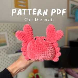 Puede incluir: Un cangrejo de peluche de color coral con pinzas en forma de corazón y ojos negros. El texto "PATTERN PDF" y "Carl the crab" se muestran encima del cangrejo. El cangrejo está hecho de un material suave y texturizado.