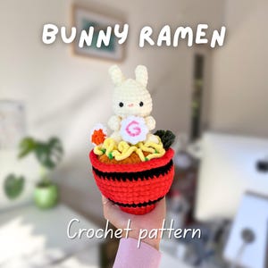 Puede incluir: Un cuenco de ramen tejido a ganchillo con una figura de conejo encima. El cuenco rojo, con rayas negras, está lleno de fideos amarillos y coronado con un conejo blanco, huevo y algas verdes. El texto dice "BUNNY RAMEN" y "Crochet pattern".