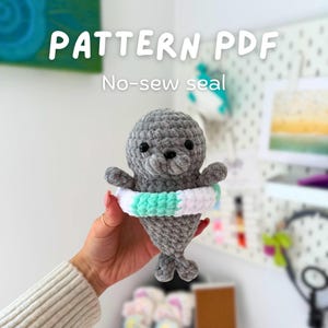 Puede incluir: Un juguete de foca gris tejido a ganchillo con un salvavidas blanco y verde menta. El texto "PATTERN PDF" y "No-sew seal" se muestran en blanco. La foca tiene ojos negros y aletas pequeñas.
