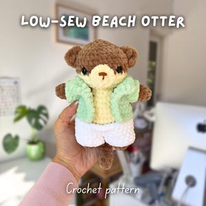 Puede incluir: Un peluche de nutria de ganchillo con una chaqueta verde claro y pantalones cortos blancos. La nutria es marrón con una barriga amarilla. El texto "LOW-SEW BEACH OTTER" y "Crochet pattern" son visibles.
