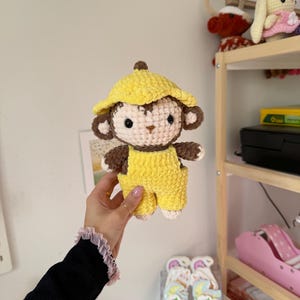 May include: A handmade crochet monkey doll wearing a yellow hat and overalls. The monkey has brown and cream-colored details, including its face, ears, and hands. The doll is held up in front of a white wall.