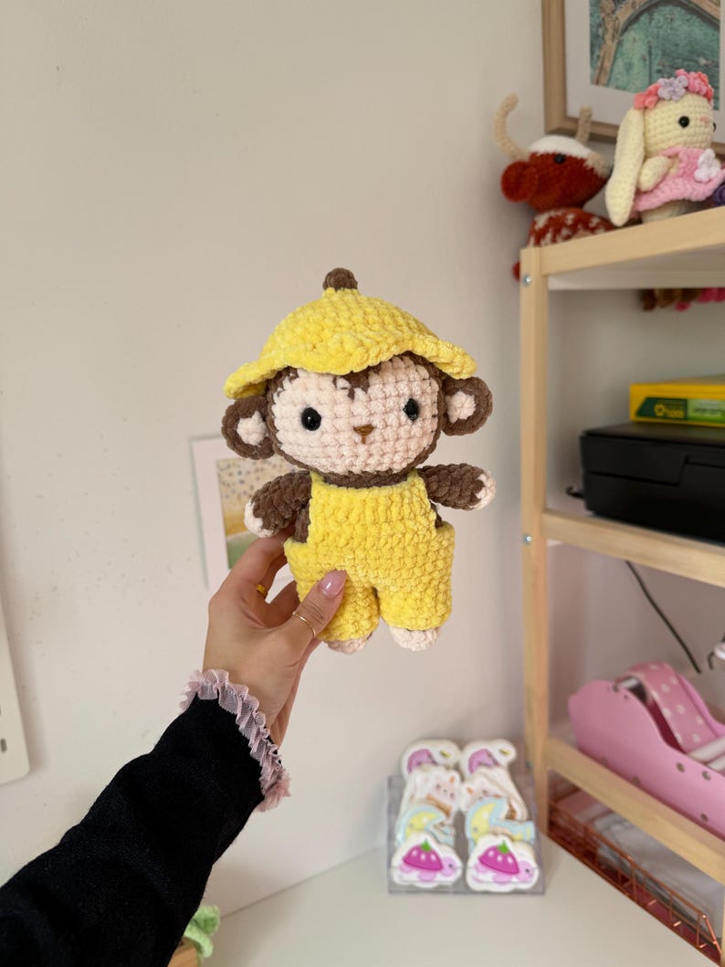 May include: A handmade crochet monkey doll wearing a yellow hat and overalls. The monkey has brown and cream-colored details, including its face, ears, and hands. The doll is held up in front of a white wall.