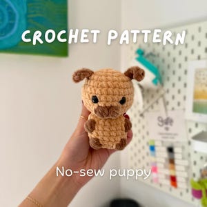 Puede incluir: Un cachorro de ganchillo en tonos marrón y beige, con ojos negros y orejas pequeñas. Se muestra el texto "CROCHET PATTERN" y "No-sew puppy". El cachorro está en una mano.