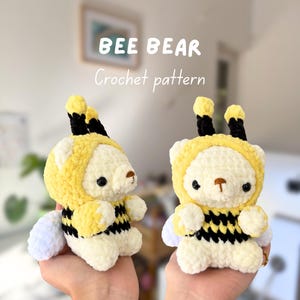 Puede incluir: Dos osos de ganchillo hechos a mano. Los osos son de color crema con detalles amarillos y negros, incluyendo una barriga a rayas de abeja y una capucha con antenas. El texto "BEE BEAR Crochet pattern" se muestra encima de los osos.
