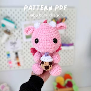 Puede incluir: Un peluche de dragón rosa hecho a mano con ganchillo, con ojos negros, cuernos blancos y pequeñas alas rosas. El dragón sostiene una taza de té boba en miniatura. El texto "PATTERN PDF" y "Baby boba dragon" son visibles.