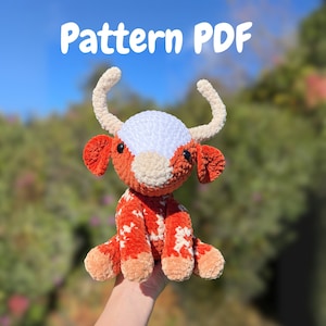 Puede incluir: Patrón de crochet PDF para una vaca de peluche con cara blanca y cuerpo naranja y blanco. La vaca sostiene una pequeña flor blanca en la boca.