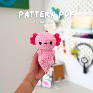 May include: A pink, no-sew axolotl plushie with bright pink gills and black eyes. The text "PATTERN PDF" and "no-sew axolotl" are displayed above the plushie. The axolotl is handmade and has a soft, textured appearance.