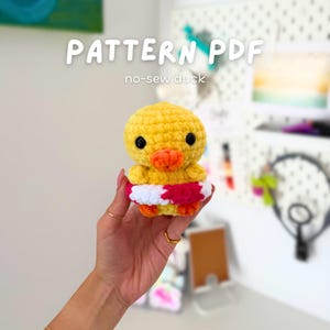 May include: A crocheted yellow duck with an orange beak and feet, wearing a red and white striped floatie. The text "PATTERN PDF" and "no-sew duck" are displayed above the duck.