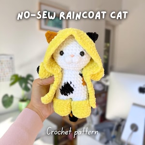 May include: A handmade crochet cat wearing a yellow raincoat. The cat is white with black and brown accents. The text "NO-SEW RAINCOAT CAT" and "Crochet pattern" are displayed on the image.