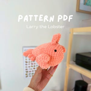 Puede incluir: Un juguete de langosta de ganchillo de color coral con ojos negros y una superficie texturizada. El texto "PATTERN PDF" y "Larry the Lobster" se muestran encima del juguete. El juguete se sostiene en una mano.