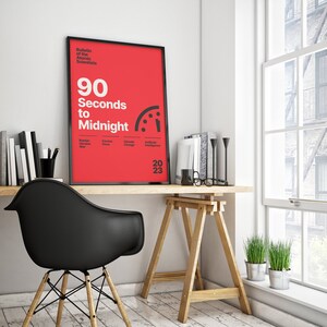 May include: A black framed poster with a red background and white text that reads "90 Seconds to Midnight" with a clock face graphic and the year "2023". The poster is hanging on a white wall above a wooden desk with a black chair and a wooden easel.