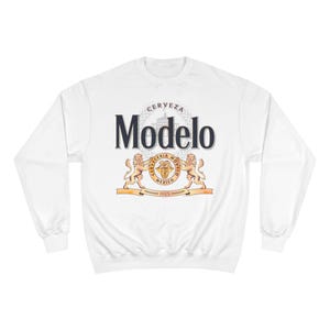 Op de afbeelding: Witte sweatshirt met het Modelo-logo. Het ontwerp bevat het woord "Modelo" in marineblauw, een wapen met de woorden "CERVEZA" en "MEXICO" en twee gouden leeuwen. Het jaar "1925" is ook zichtbaar.