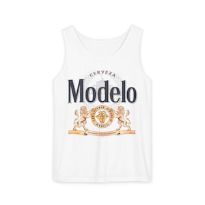 May include: White tank top featuring the Modelo Cerveza logo. The logo includes the word "Modelo" in navy blue, a crest with two lions, and the text "Cerveceria Modelo Mexico 1925".