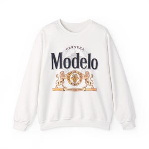 Op de afbeelding: Witte crewneck sweatshirt met het Modelo bierlogo. Het logo bevat het woord "Modelo" in marineblauw, een wapen met twee leeuwen en de tekst "CERVECERIA MODELO MEXICO 1925".