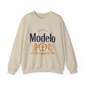 Op de afbeelding: Een beige crewneck sweatshirt met het "Modelo" bierlogo. Het logo bevat het woord "Modelo" in marineblauw, het woord "CERVEZA" erboven en een wapen met leeuwen en de woorden "CERVECERIA MODELO MEXICO 1925" eronder. De sweatshirt heeft een neutrale kleur.