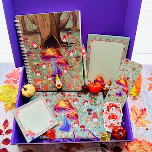 May include: A purple box filled with stationery items featuring a whimsical mushroom and forest theme. Includes a spiral notebook, notepad, pen, bookmark, and stickers. Decorative pumpkins and fall leaves surround the box, creating a seasonal aesthetic.