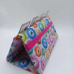May include: A fabric bingo card holder with a gray background and colorful bingo ball patterns. The holder has a bright pink elastic band and holds bingo cards. The bingo balls are in various colors, including blue, orange, and pink.