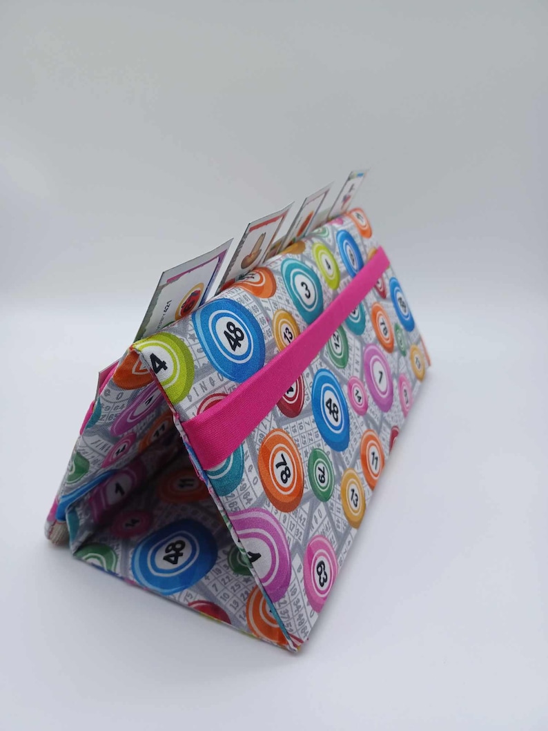 May include: A fabric bingo card holder with a gray background and colorful bingo ball patterns. The holder has a bright pink elastic band and holds bingo cards. The bingo balls are in various colors, including blue, orange, and pink.