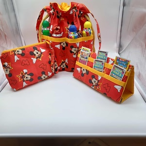 May include: A set of red fabric accessories featuring a Mickey Mouse pattern. The set includes a drawstring bag, a zippered pouch, and a card holder. The bag has a yellow interior and gold rope handles. The card holder displays several cards.