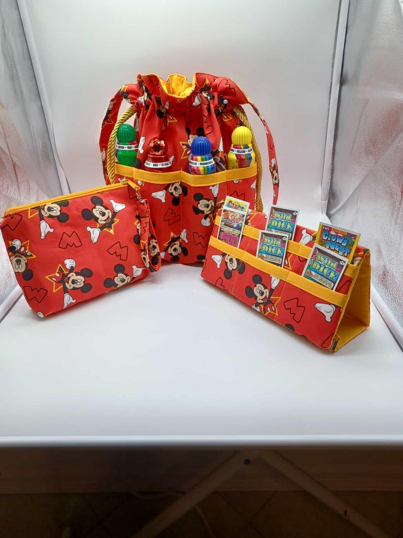 May include: A set of red fabric accessories featuring a Mickey Mouse pattern. The set includes a drawstring bag, a zippered pouch, and a card holder. The bag has a yellow interior and gold rope handles. The card holder displays several cards.