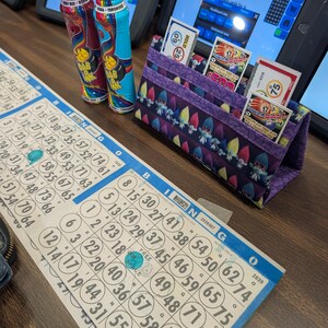 May include: A bingo game setup featuring two Dab Ink markers, a purple fabric card holder with bingo cards, and a bingo card with blue and white markings. The markers are red and turquoise. The card holder has a whimsical gnome pattern.
