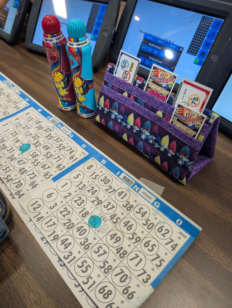 May include: A bingo game setup featuring two Dab Ink markers, a purple fabric card holder with bingo cards, and a bingo card with blue and white markings. The markers are red and turquoise. The card holder has a whimsical gnome pattern.
