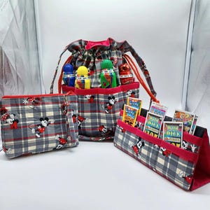 May include: A set of three matching fabric bags with a gray plaid pattern and Mickey Mouse illustrations. The set includes a drawstring bag, a zippered pouch, and a stand-up organizer with red trim. The drawstring bag holds several small bottles.