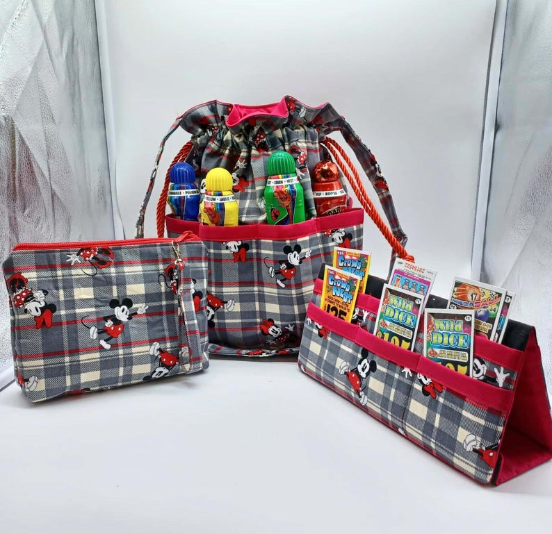 May include: A set of three matching fabric bags with a gray plaid pattern and Mickey Mouse illustrations. The set includes a drawstring bag, a zippered pouch, and a stand-up organizer with red trim. The drawstring bag holds several small bottles.