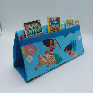 May include: A blue fabric organizer with a Moana theme, featuring the character and tropical flowers. The organizer holds small game packs labeled "Wild Dice", "Crows Nest", and "Shark Attack". The organizer has a blue trim.