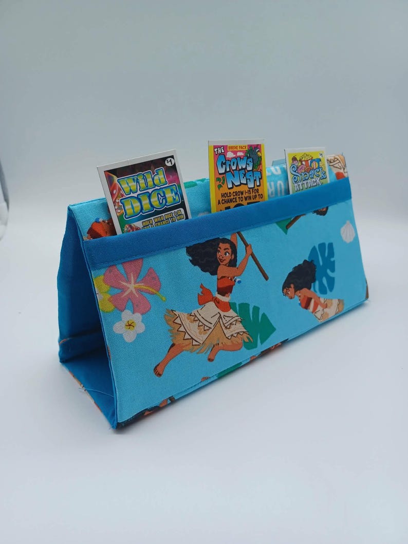 May include: A blue fabric organizer with a Moana theme, featuring the character and tropical flowers. The organizer holds small game packs labeled "Wild Dice", "Crows Nest", and "Shark Attack". The organizer has a blue trim.