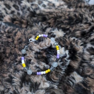 Op de afbeelding: Een kralenarmband met de kleuren van de non-binaire pride-vlag. De armband heeft gele, witte, paarse en zwarte kralen, met daartussen heldere, ronde kralen. De sluiting is zilverkleurig.