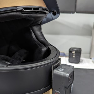 May include: A black motorcycle helmet with a gold top section, featuring a GoPro camera attached to the side. The camera is black with a red record button. The helmet has a dark visor and a sticker.