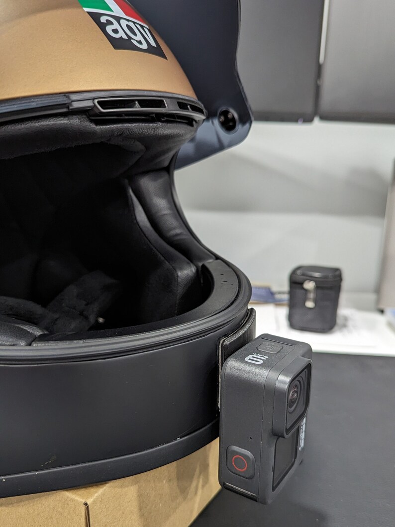 May include: A black motorcycle helmet with a gold top section, featuring a GoPro camera attached to the side. The camera is black with a red record button. The helmet has a dark visor and a sticker.