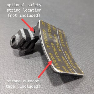 May include: A black plastic mount with a textured finish, featuring a mounting bracket and a curved adhesive pad. The text "optional safety string location (not included)" and "strong outdoor tape (included)" are visible.
