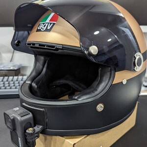 May include: A black and gold motorcycle helmet with a GoPro camera attached. The helmet has a visor and the AGV logo. The helmet is sitting on a cardboard box.