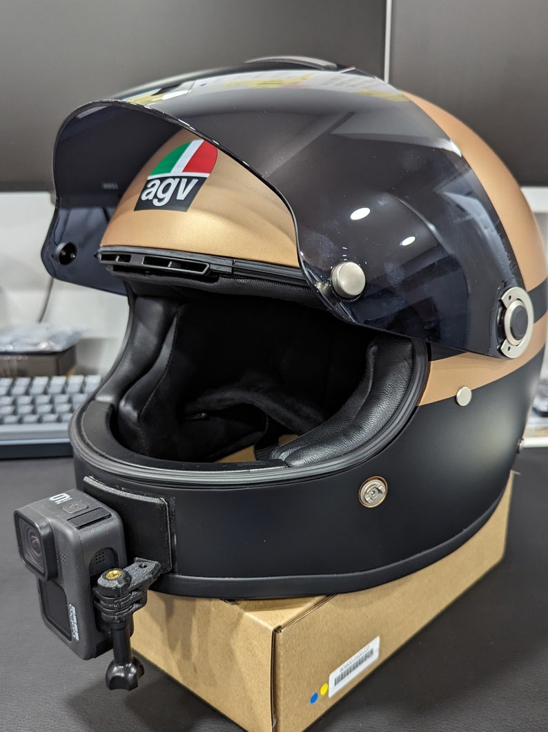 May include: A black and gold motorcycle helmet with a GoPro camera attached. The helmet has a visor and the AGV logo. The helmet is sitting on a cardboard box.