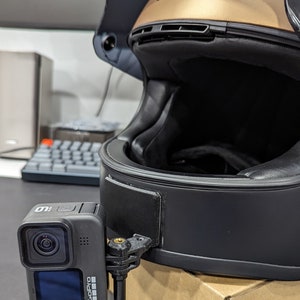 May include: A black motorcycle helmet with a gold top and a GoPro camera attached. The camera is mounted on the side of the helmet with a black bracket. The helmet has a black interior and a clear visor.