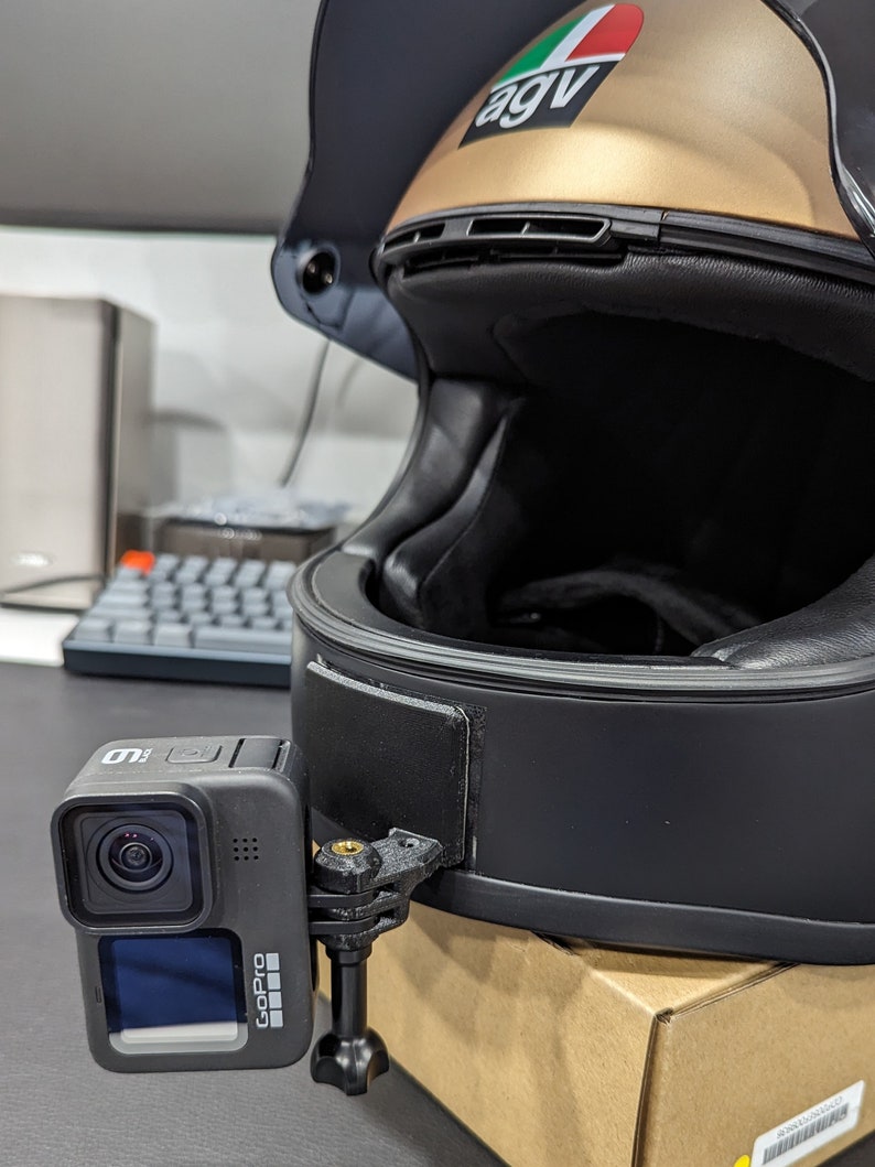 May include: A black motorcycle helmet with a gold top and a GoPro camera attached. The camera is mounted on the side of the helmet with a black bracket. The helmet has a black interior and a clear visor.