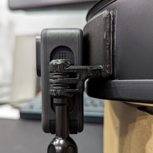May include: A black action camera is mounted on a black bracket attached to a black object. The camera is rectangular with a lens and several buttons. The bracket appears to be 3D-printed. The object is round and has a smooth, matte finish.