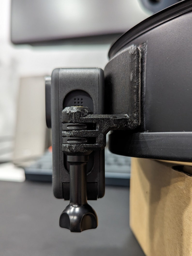 May include: A black action camera is mounted on a black bracket attached to a black object. The camera is rectangular with a lens and several buttons. The bracket appears to be 3D-printed. The object is round and has a smooth, matte finish.