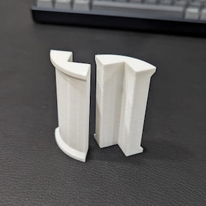 May include: Two white plastic pieces, each with a curved shape and a rectangular opening. The pieces are designed to fit together to create a larger, more complex shape.