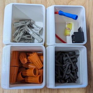 May include: Four white plastic containers with different hardware items. The top left container has a pile of gray plastic anchors. The top right container has a blue wire, two yellow connectors, and a black electrical component. The bottom left container has a pile of orange wire connectors. The bottom right container has a pile of black screws.