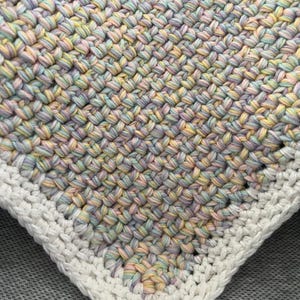 May include: A crocheted baby blanket with a white border. The blanket is made with a multi-coloured yarn in shades of pink, blue, and yellow.