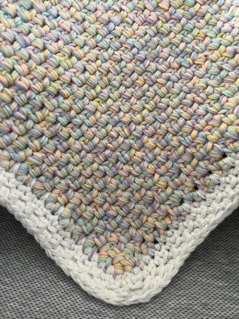 May include: A crocheted baby blanket with a white border. The blanket is made with a multi-coloured yarn in shades of pink, blue, and yellow.