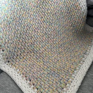 May include: A crocheted baby blanket with a white border. The blanket is made with a multi-coloured yarn in shades of pink, blue, and yellow.