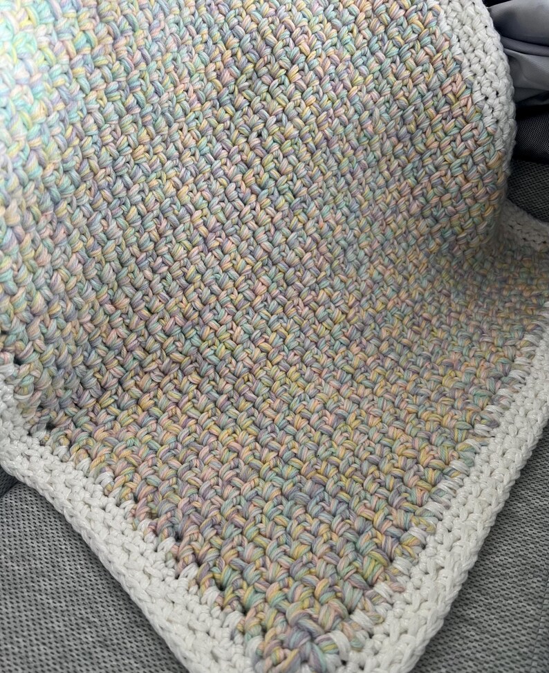 May include: A crocheted baby blanket with a white border. The blanket is made with a multi-coloured yarn in shades of pink, blue, and yellow.