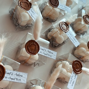 May include: A collection of small, clear boxes filled with white, round, scented candles. Each box has a gold wax seal with a wreath design and a handwritten tag with the text "Donut & Andreia 19 August 2023".