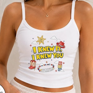 May include: A white ribbed tank top featuring a graphic print with the text I KNEW IT, I KNEW YOU. The design includes a yellow sheriff star, a red cowboy hat, red cowboy boots, a beaded bracelet, and a stack of colorful alphabet blocks.