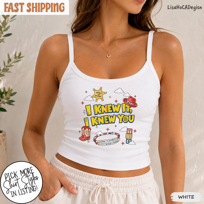 May include: A white ribbed tank top featuring a graphic print with the text I KNEW IT, I KNEW YOU. The design includes a yellow sheriff star, a red cowboy hat, red cowboy boots, a beaded bracelet, and a stack of colorful alphabet blocks.